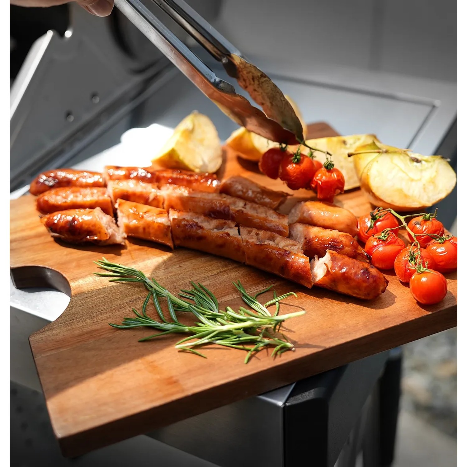 Angeschnittene Bratwurst, Tomaten, Apfel und Rosmarin auf Schneidebrett auf Taino Gasgrill.