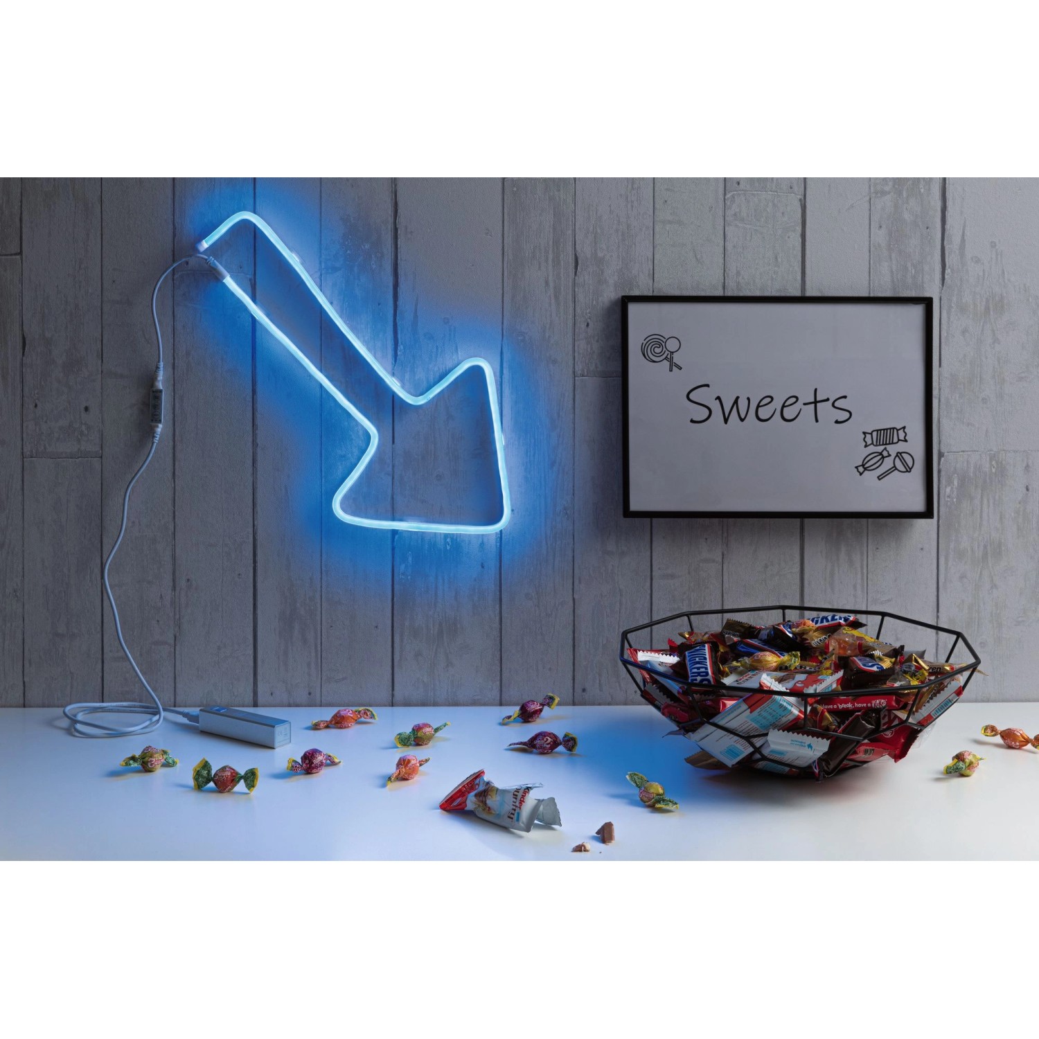 Paulmann Neon Colorflex LED-Streifen als blauer Pfeil an Wand, Süßigkeiten im Vordergrund.
