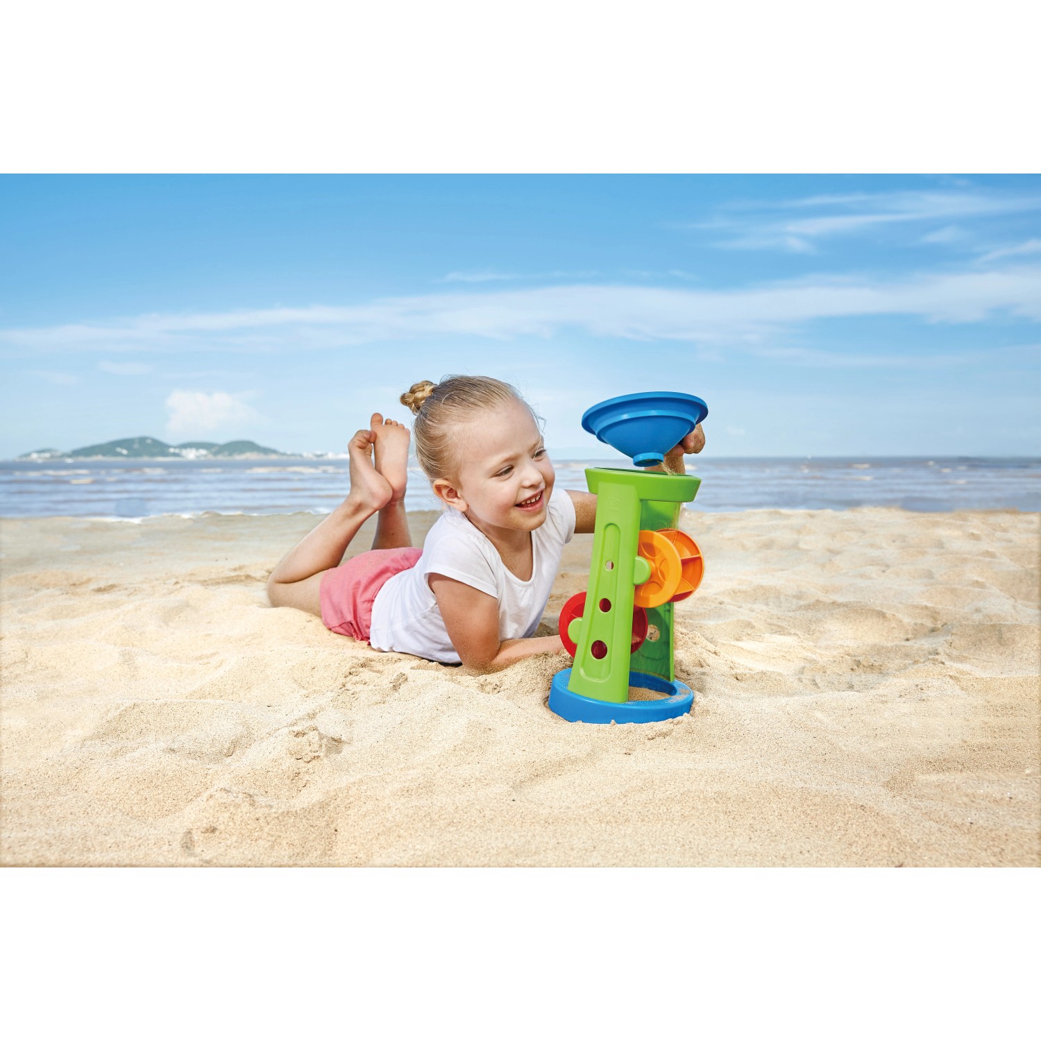 Kind spielt mit Hape Sandspielzeug-Set (Sandmühle, Sieb) am Strand. Mehrfarbiges Kunststoff-Spielzeug.