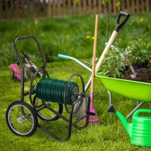 Brauner Relaxdays Gartenschlauchwagen aus Metall mit grünem Schlauch, Kurbel, Rädern und Zubehörkorb im Garten.