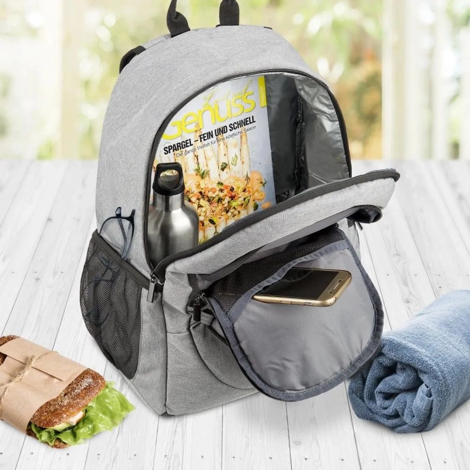 Grauer Bomoe IceBreezer Kühlrucksack (25L) mit Zubehör wie Getränk, Zeitschrift und Sandwich.
