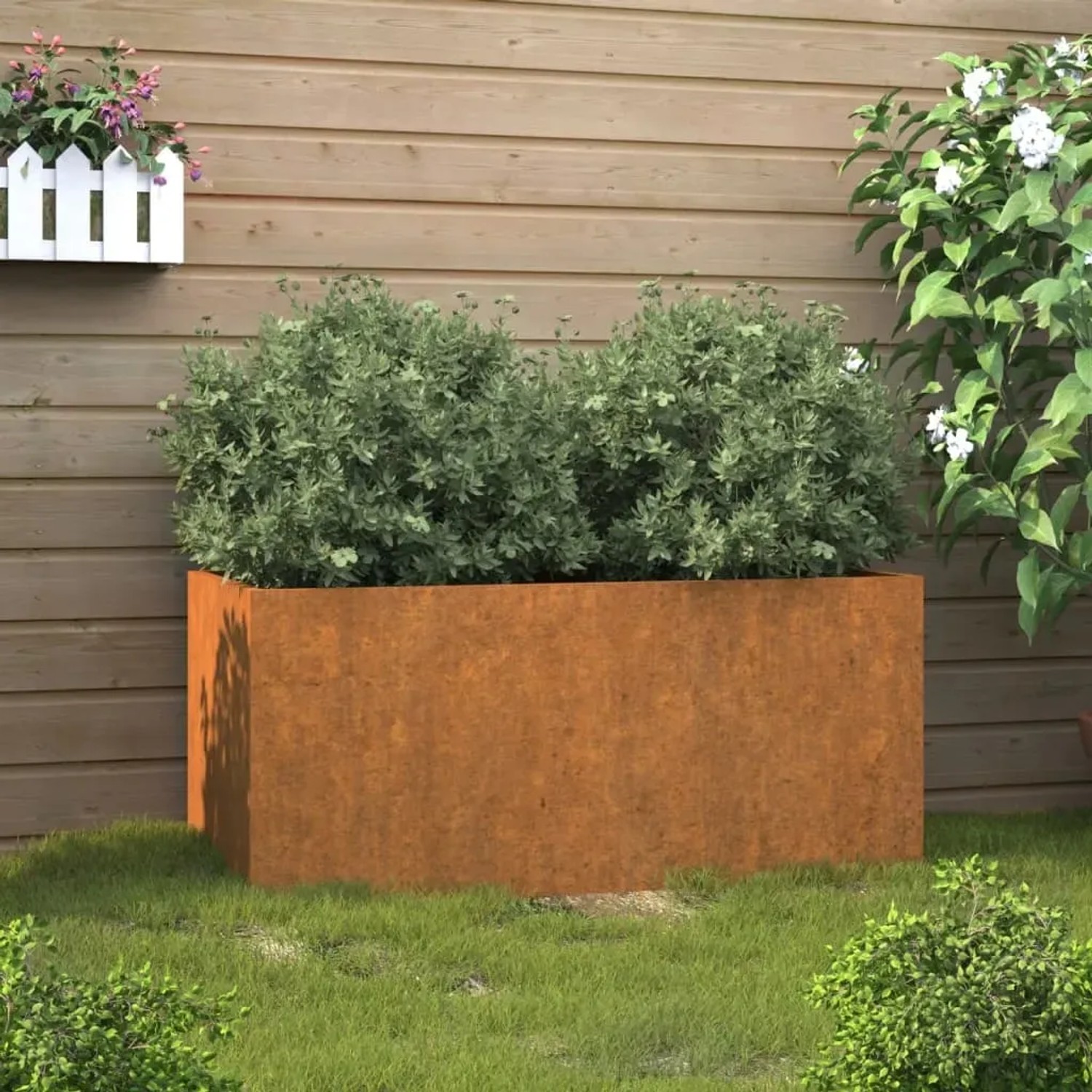 Rechteckiger Pflanzkübel aus Cortenstahl, rostbraun, mit grünen Pflanzen im Garten. Maße: 62x30x29 cm.