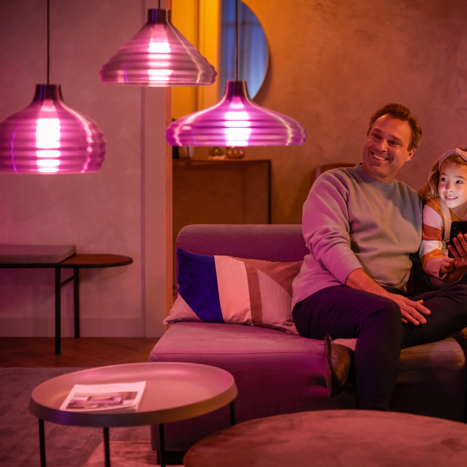 Szene mit Philips Hue Leuchten: Vater und Tochter auf Sofa unter smarten Hängelampen.