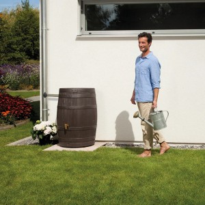 4Rain Regenfass Vino 250 l in Dunkelbraun mit Holzoptik im Garten, daneben eine Person mit Gießkanne.