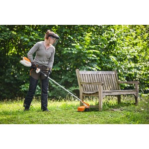 Frau mit Gesichtsschutz trimmt Rasen mit orange-grauer Stihl Akku-Motorsense FSA 86 R neben Holzbank im Garten.