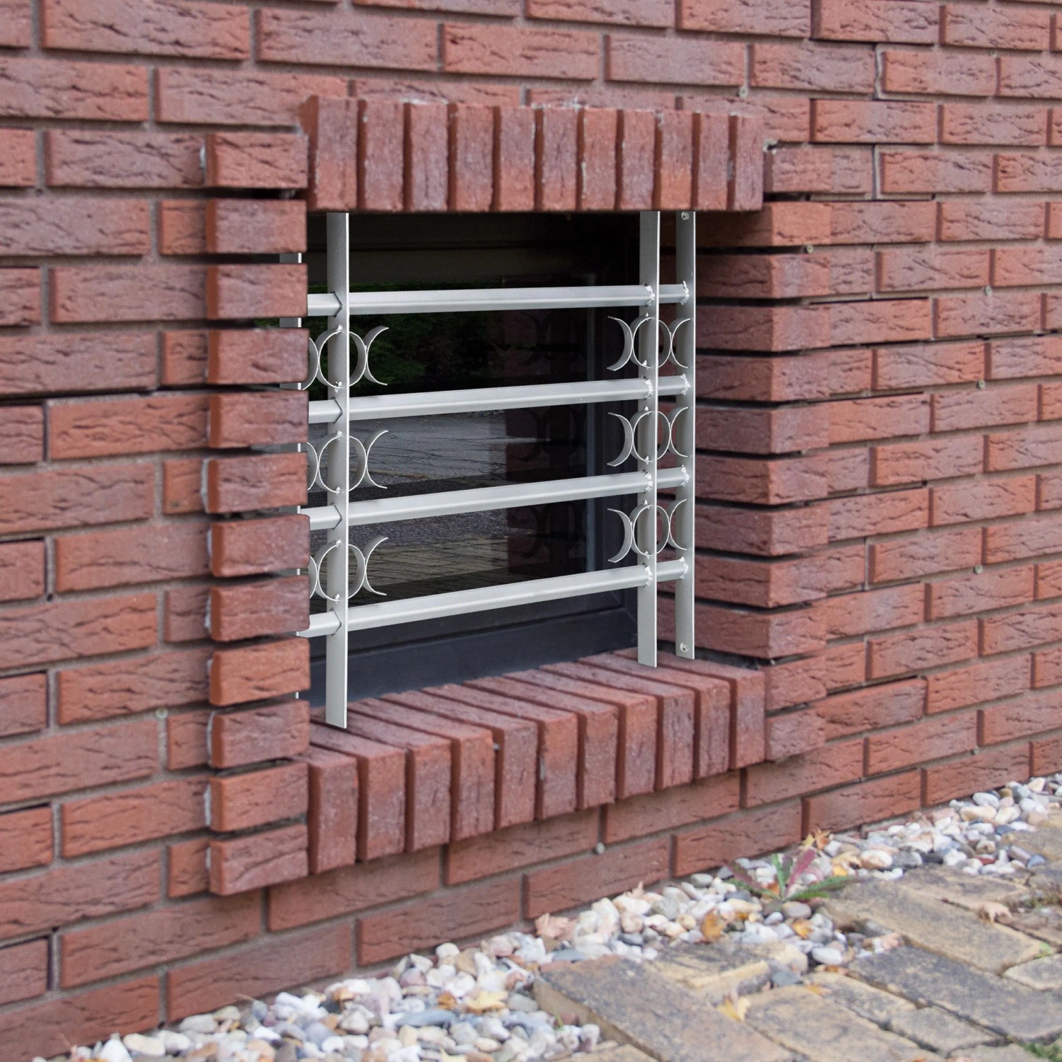 Graues, ausziehbares Stahl-Fenstergitter mit Zierelementen als Einbruchschutz an Ziegelwand. Für Kellerfenster.