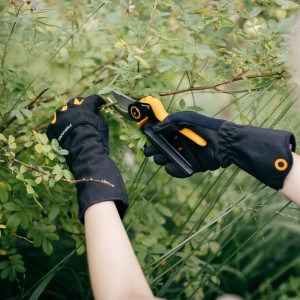 Hände mit Fiskars Strauchpflege Handschuhen (Größe 8, Schwarz-Orange) schneiden Strauch.