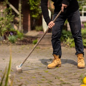 Mann reinigt mit Drahtbesen aus Buchenholz und Stahl Pflastersteine von Unkraut.
