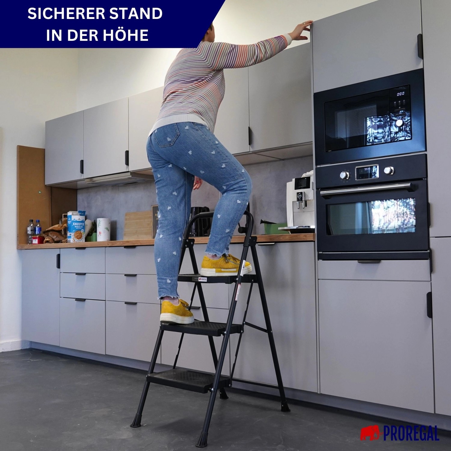 Frau auf PROREGAL Trittleiter (3 Stufen) in Küche, um an Schrank zu gelangen. Klappbare Haushaltsleiter aus Stahl, schwarz.