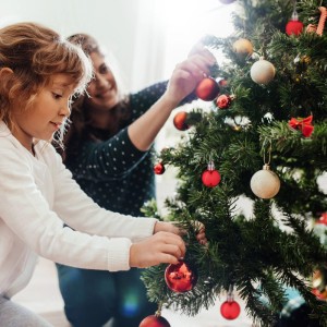 Ein Kind und ein Erwachsener schmücken einen Weihnachtsbaum mit glänzenden Weihnachtskugeln. 96er Set Christbaumschmuck aus Kunststoff.