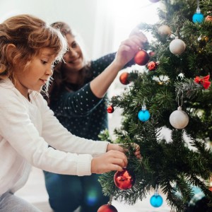 Kind und Erwachsene schmücken einen Tannenbaum mit glänzenden roten, blauen und hellen Weihnachtskugeln als Christbaumschmuck.