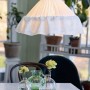 Weiße PR Home Stina Pendelleuchte aus Baumwollstoff mit Plisseefalten und Rüschensaum über einem Tisch mit Blumen.