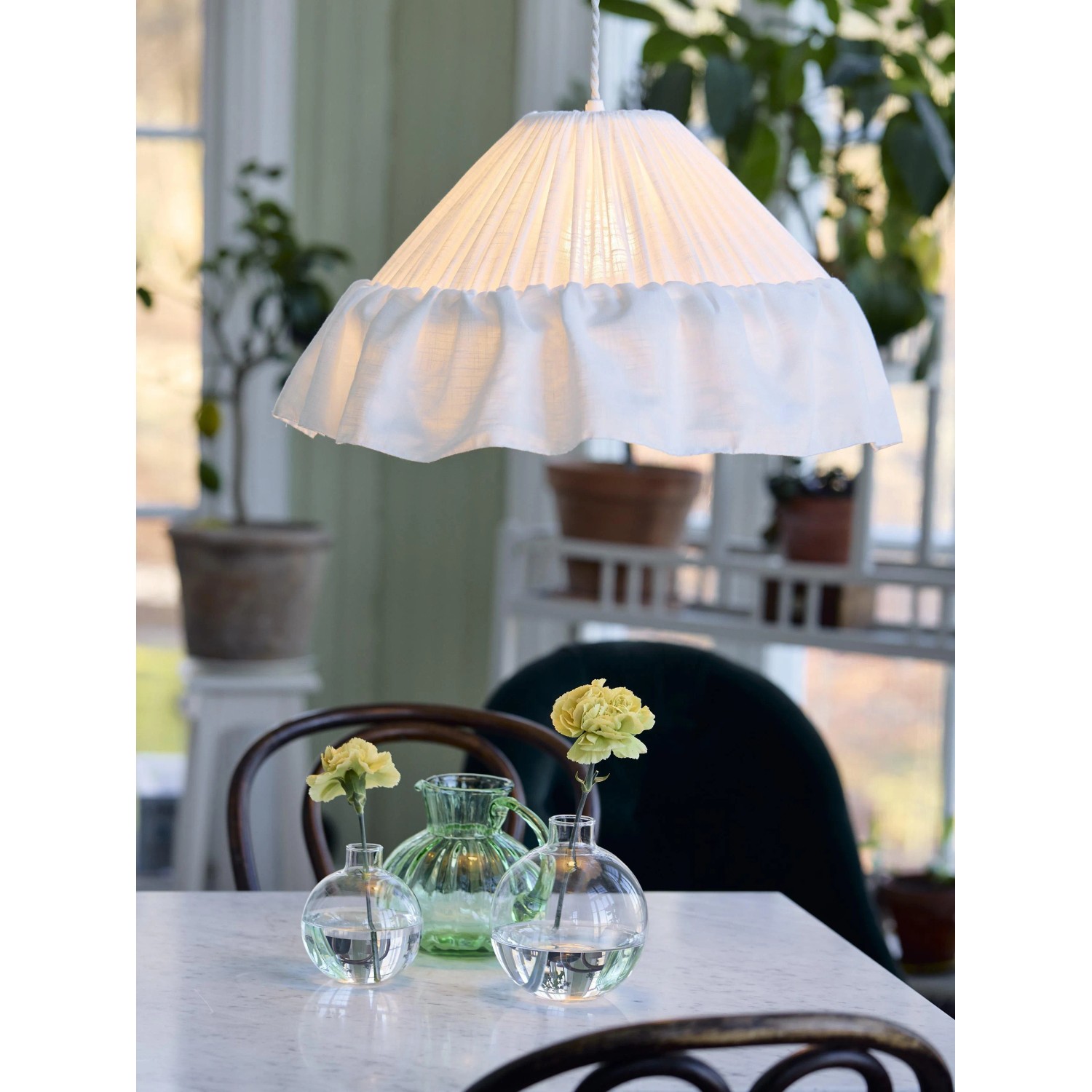Weiße PR Home Stina Pendelleuchte aus Baumwollstoff mit Plisseefalten und Rüschensaum über einem Tisch mit Blumen.