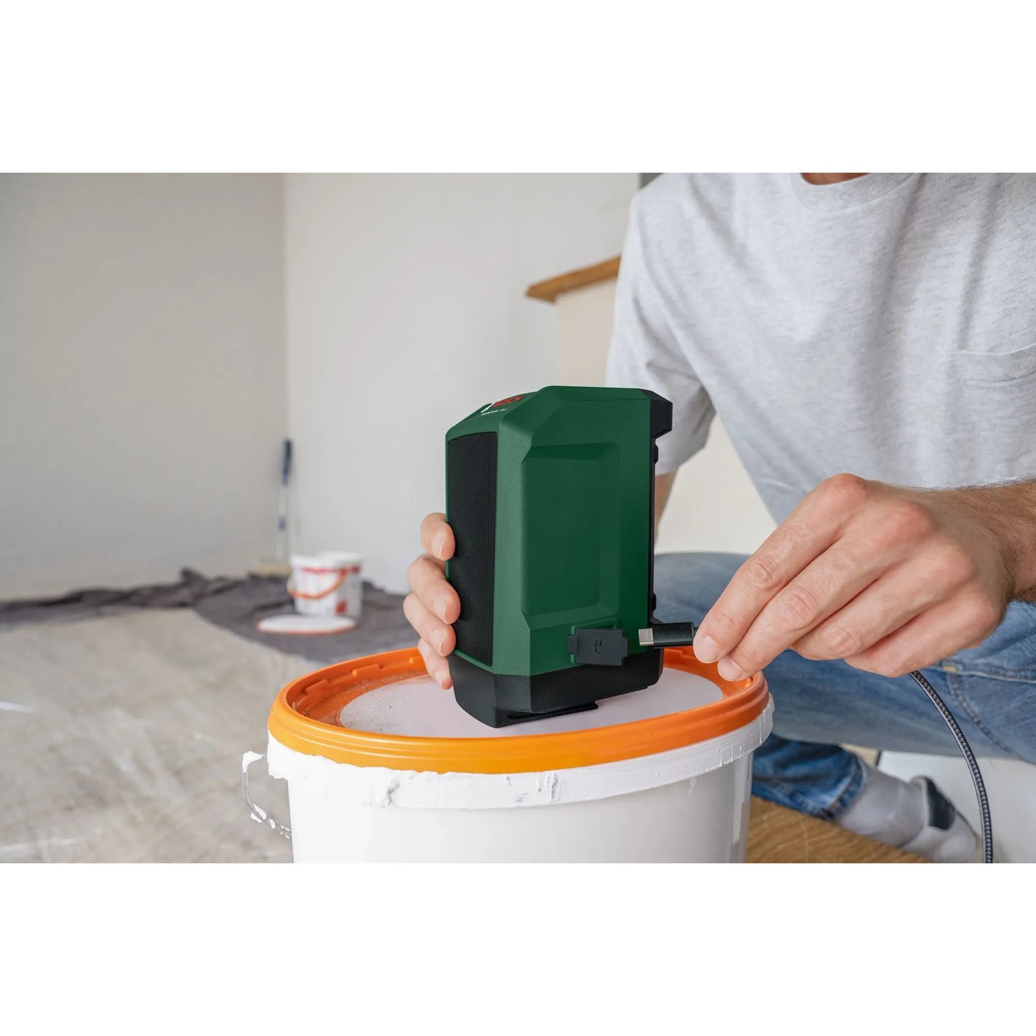 Bosch UniversalSpeaker 18V-1 Akku-Bluetooth-Box in Grün, per USB-C verbunden, auf Farbeimer in Arbeitsumgebung.