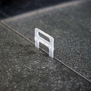 Transparente Xpotool Zuglasche für 1,0 mm Fugen im Fliesen Nivelliersystem, eingesetzt zwischen dunklen Fliesen. Verlegehilfe.