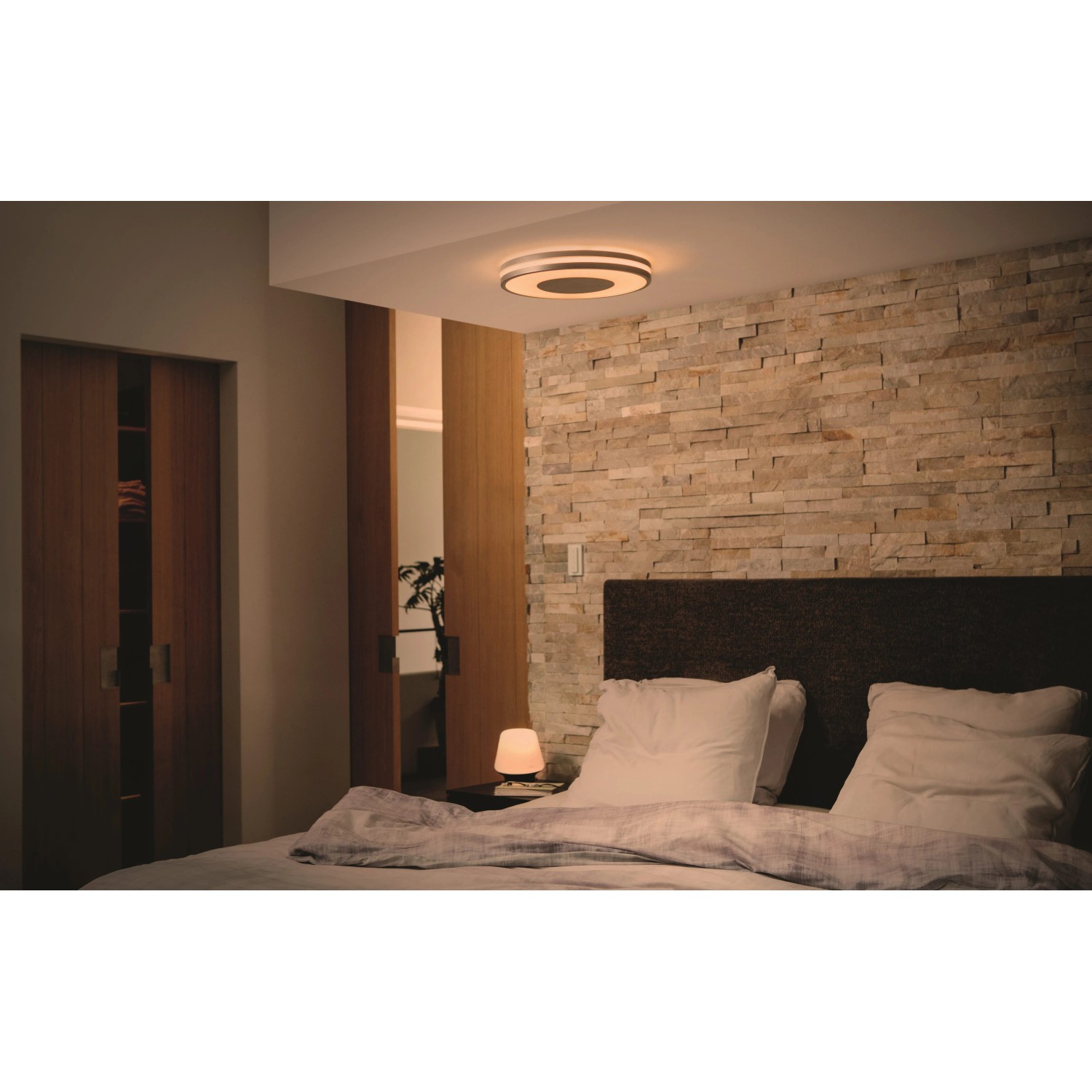 Philips Hue Being Deckenleuchte in Silber, montiert in einem Schlafzimmer mit Steinwand.
