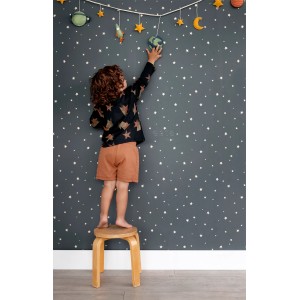 Kind auf Hocker vor dunkelblauer ESTAhome Glow In The Dark Tapete mit weißen Sternen. Ideal für Kinderzimmer.