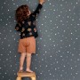 Kind auf Hocker vor dunkelblauer ESTAhome Glow In The Dark Tapete mit weißen Sternen. Ideal für Kinderzimmer.