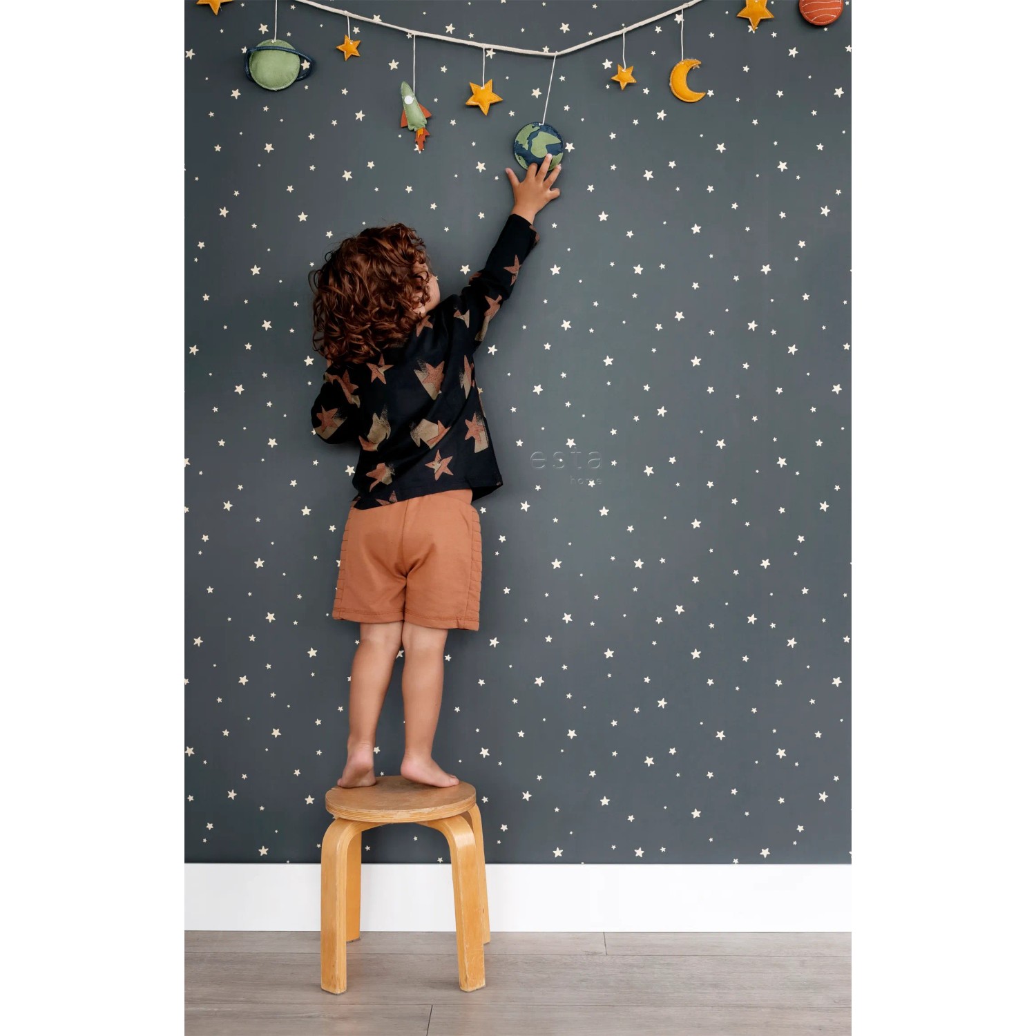 Kind auf Hocker vor dunkelblauer ESTAhome Glow In The Dark Tapete mit weißen Sternen. Ideal für Kinderzimmer.