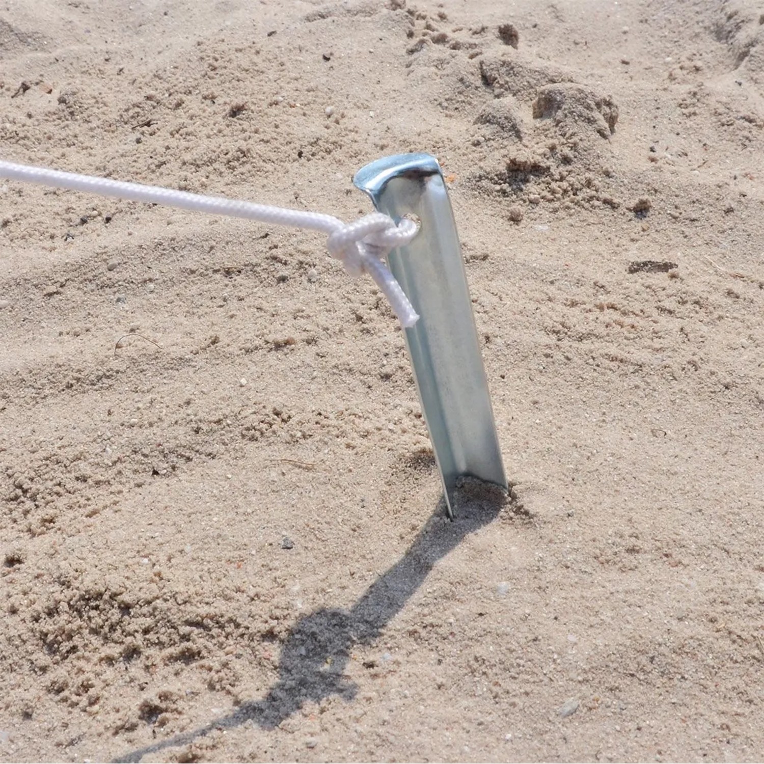 Silberfarbener V-förmiger Zelt-Hering aus verzinktem Stahl im Sand, mit weißem Seil befestigt. Für sicheren Halt beim Camping.