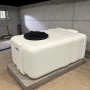 Weißer, rechteckiger KMND 200L Trinkwasser Wassertank aus PE mit schwarzem Deckel auf Betonplatte in einem Kellerraum.