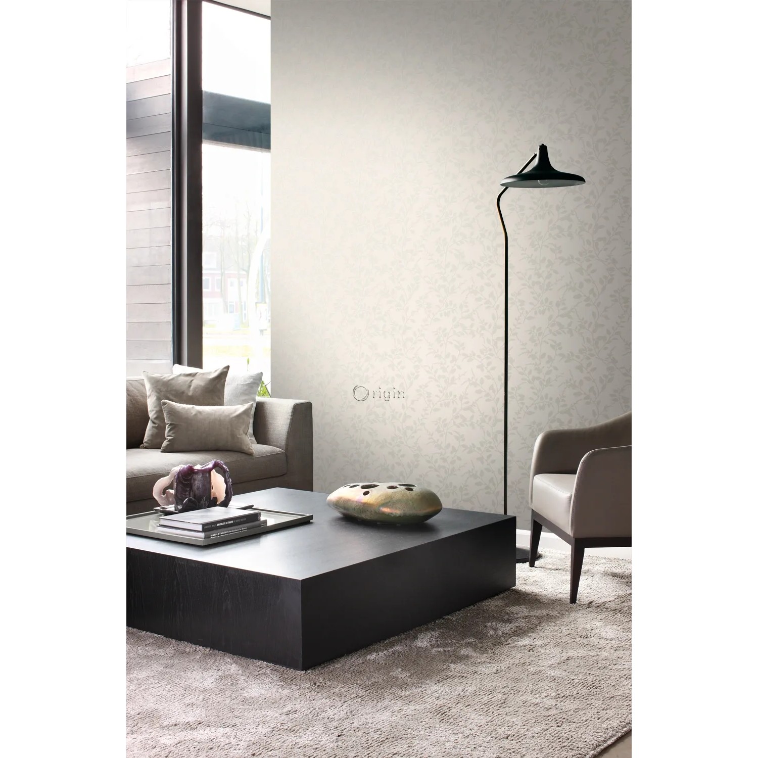 Wohnraum mit beiger Origin Wallcoverings Vliestapete Blätter-Motiv, kombiniert mit Möbeln in Braun- und Grautönen.