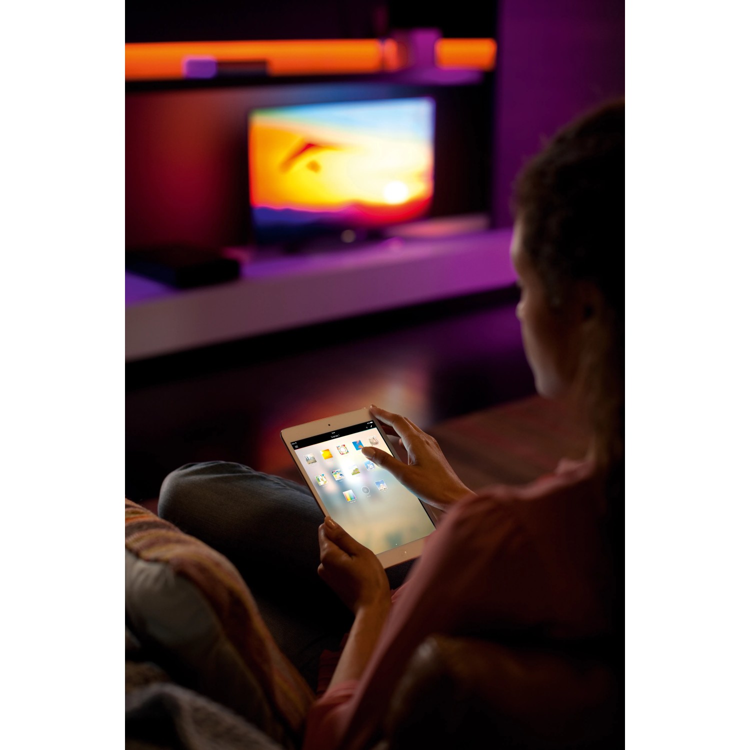 Frau steuert Philips Hue Lightstrip Plus mit Tablet, indirekte Beleuchtung im Wohnzimmer.