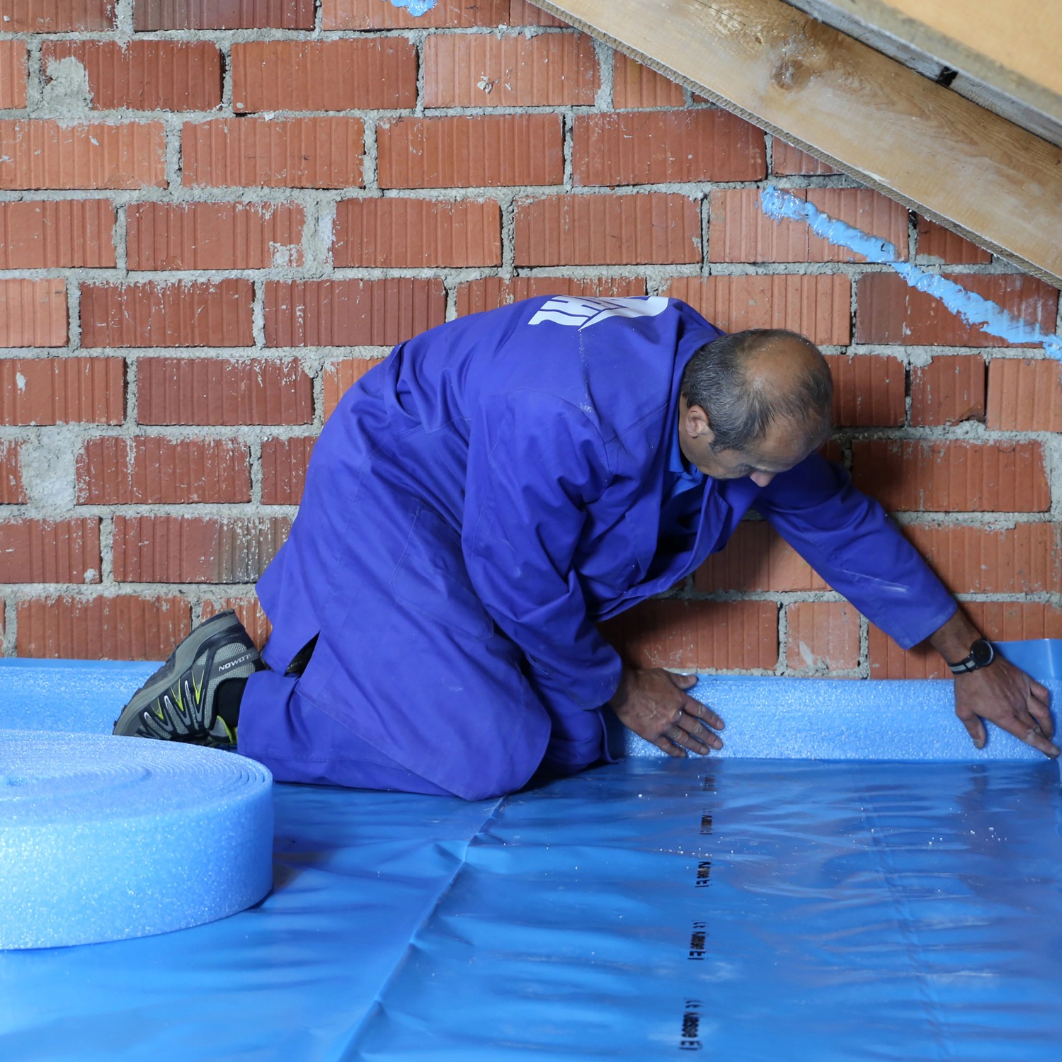 Mann verlegt blaue EPS Dachboden-Dämmplatten (120mm) auf einem Dachboden mit Ziegelwand.