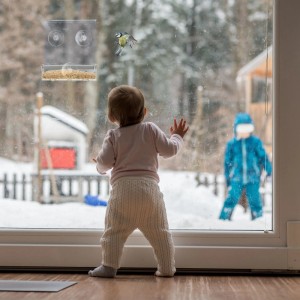 Baby beobachtet Meise am transparenten Acryl Vogelfutterhaus mit Saugnäpfen am Fenster. Winterlandschaft.