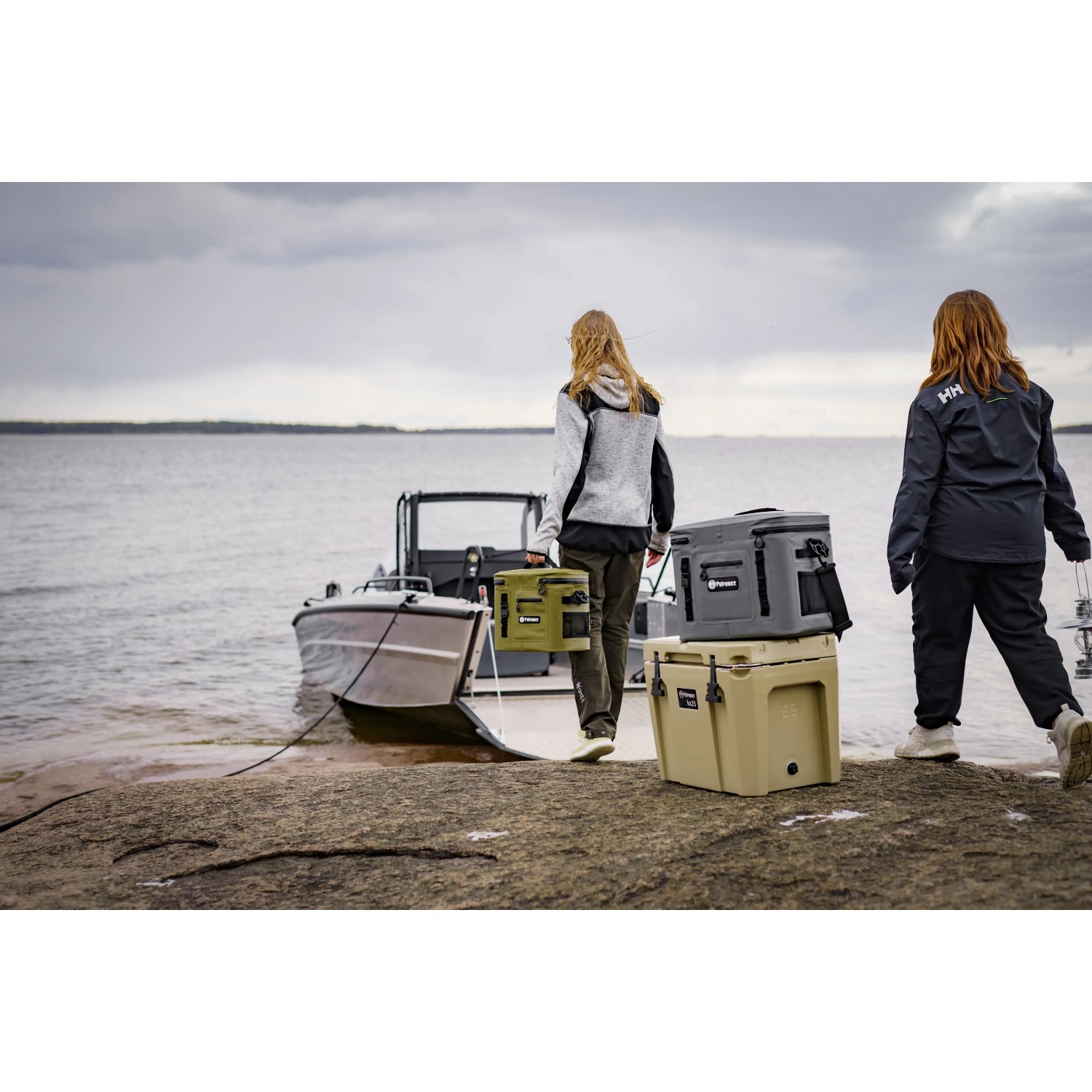 Petromax Kühlbox kx25 in Sand, gestapelt mit anderen Kühlboxen am See, transportbereit für Camping oder Ausflug.