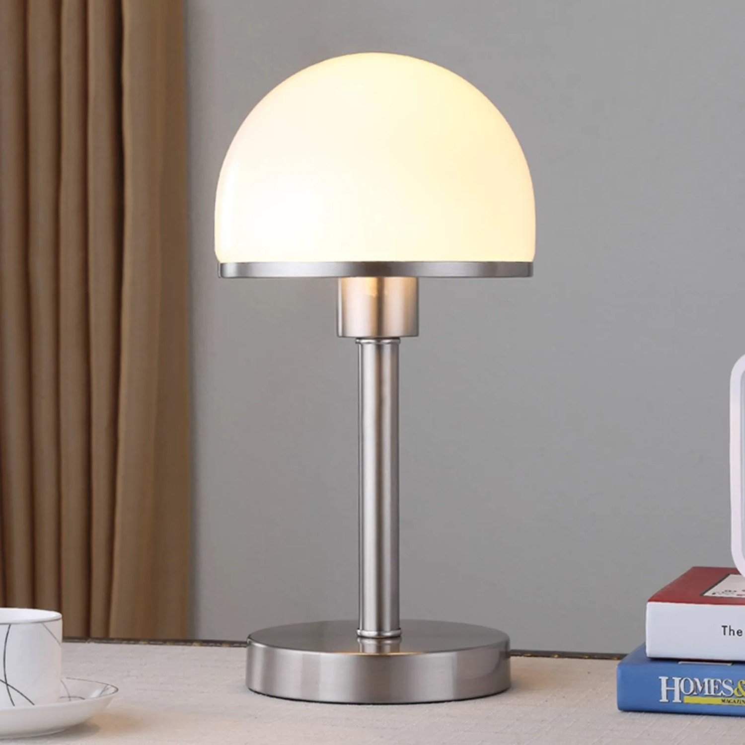 Moderne Lindby Tischlampe Jolie aus gebürstetem Metall mit weißem Halbkugelschirm, beleuchtet auf Nachttisch.
