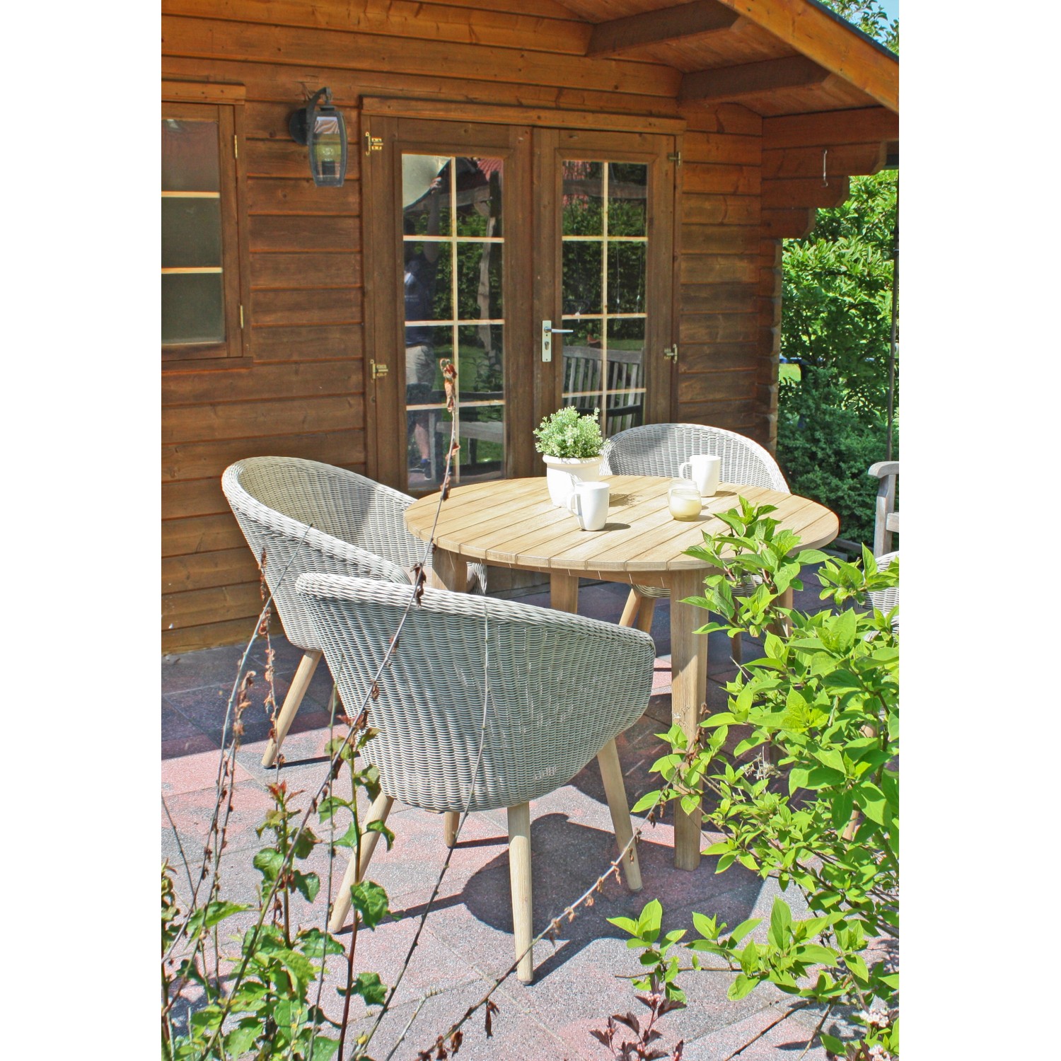 Runder Gartentisch Arvada Natur Ø 110 cm mit Stühlen auf Terrasse vor Holzhütte.
