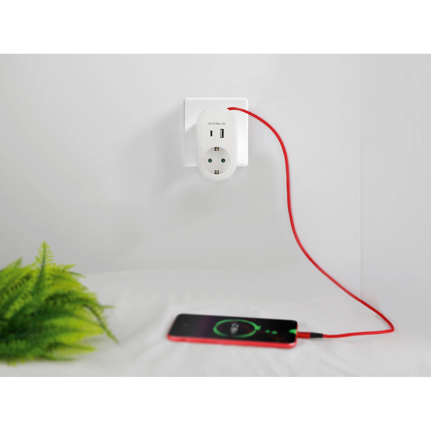 Weißer Steckdosen-Adapter mit USB-A, USB-C und Schutzkontakt-Steckdose lädt rotes Smartphone.