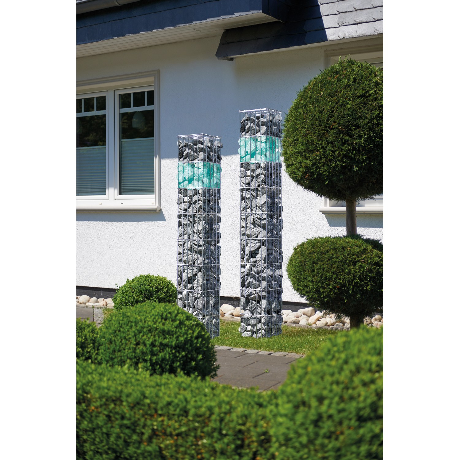 Zwei Gabionenkorb-Säulen (25x140x25 cm) im Garten, gefüllt mit grauen Steinen. Dekorative Gartengestaltung.