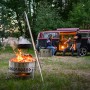 Petromax Dreibein mit Feuertopf über Lagerfeuer, im Hintergrund VW Bus mit Personen. Grillutensil für Outdoor-Küche.