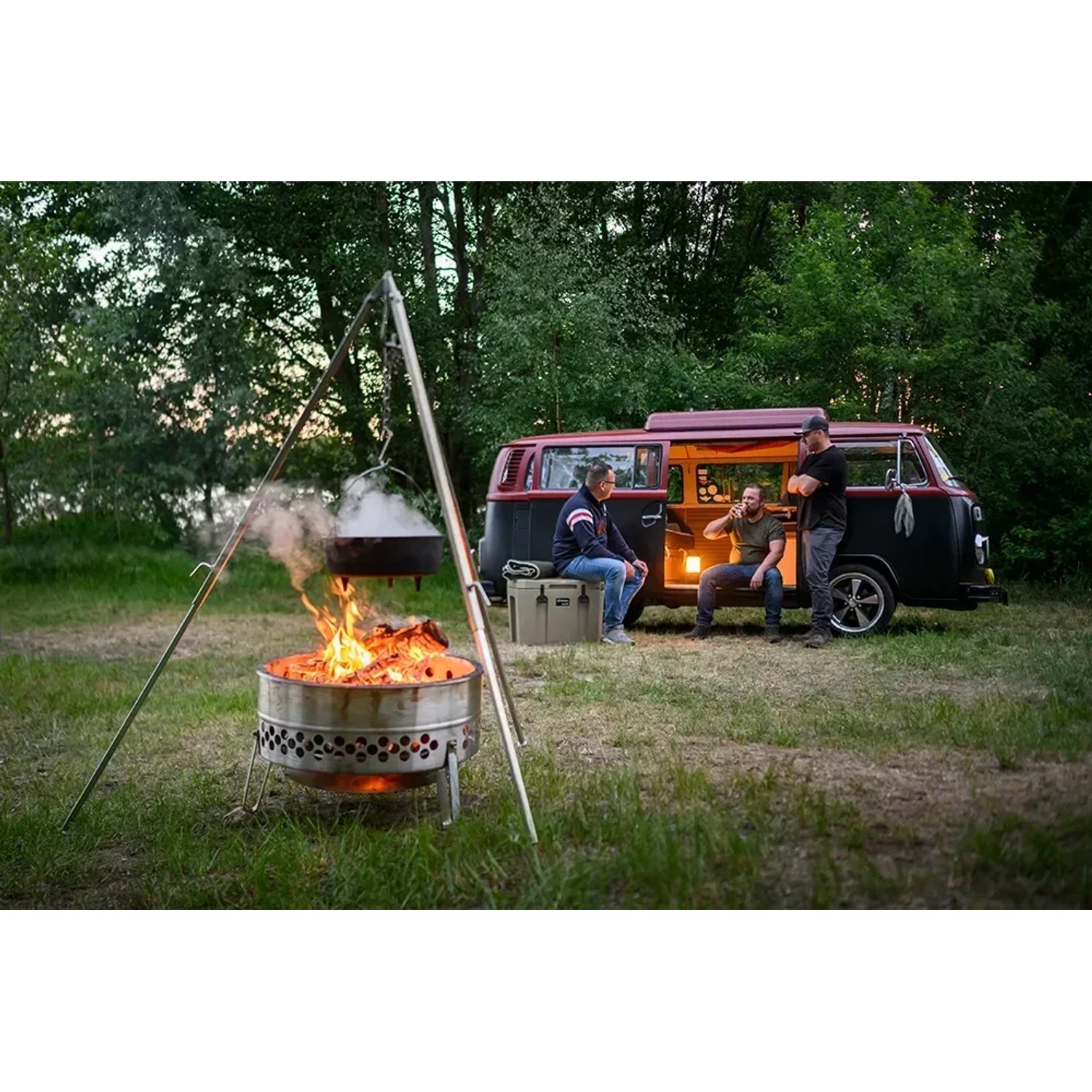Petromax Dreibein mit Feuertopf über Lagerfeuer, im Hintergrund VW Bus mit Personen. Grillutensil für Outdoor-Küche.