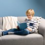 Junge liest Buch auf grauem Sofa mit Kissen und Decke vor blauer Wand.