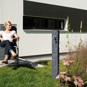 Garantia Granit Wasserzapfsäule Darkgranite mit Chromhahn im Garten, Frau sitzt daneben.