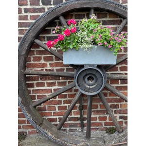 Grauer Pflanzwerk Pflanzkübel 50 cm auf Wagenrad mit bunten Blumen vor Backsteinmauer.