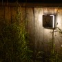 Quadratische, schwarze Lutec Solar-LED-Außenleuchte Moze an Holzwand, beleuchtet.