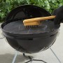 Weber Grillbürste mit Bambusgriff reinigt Grillrost. Holzkohlegrill im Hintergrund.