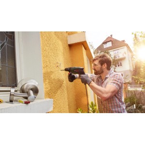 Mann bohrt mit Bosch UniversalImpact 800 Schlagbohrmaschine in gelbe Außenwand. Ideal für Heimwerker.