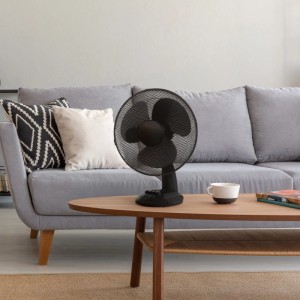 Schwarzer Tischventilator (Ø 30 cm) auf Tisch vor grauem Sofa. Ventilator mit Metallschutzgitter.