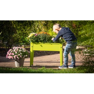 Apfelgrünes Floraworld Kinder-Hochbeet aus Metall, bepflanzt mit Blumen, Kind beugt sich darüber.