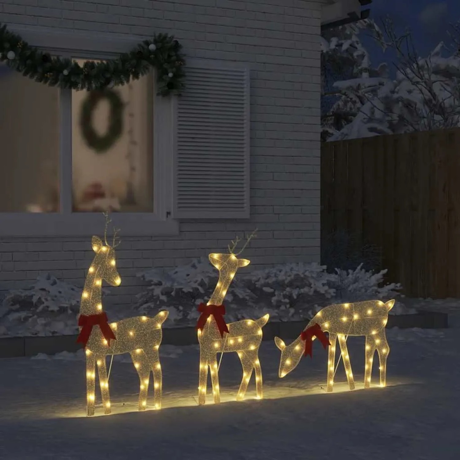 Drei goldene LED-Rentiere mit warmweißen Lichtern und roten Schleifen als Weihnachtsdekoration im Schnee vor einem Haus.