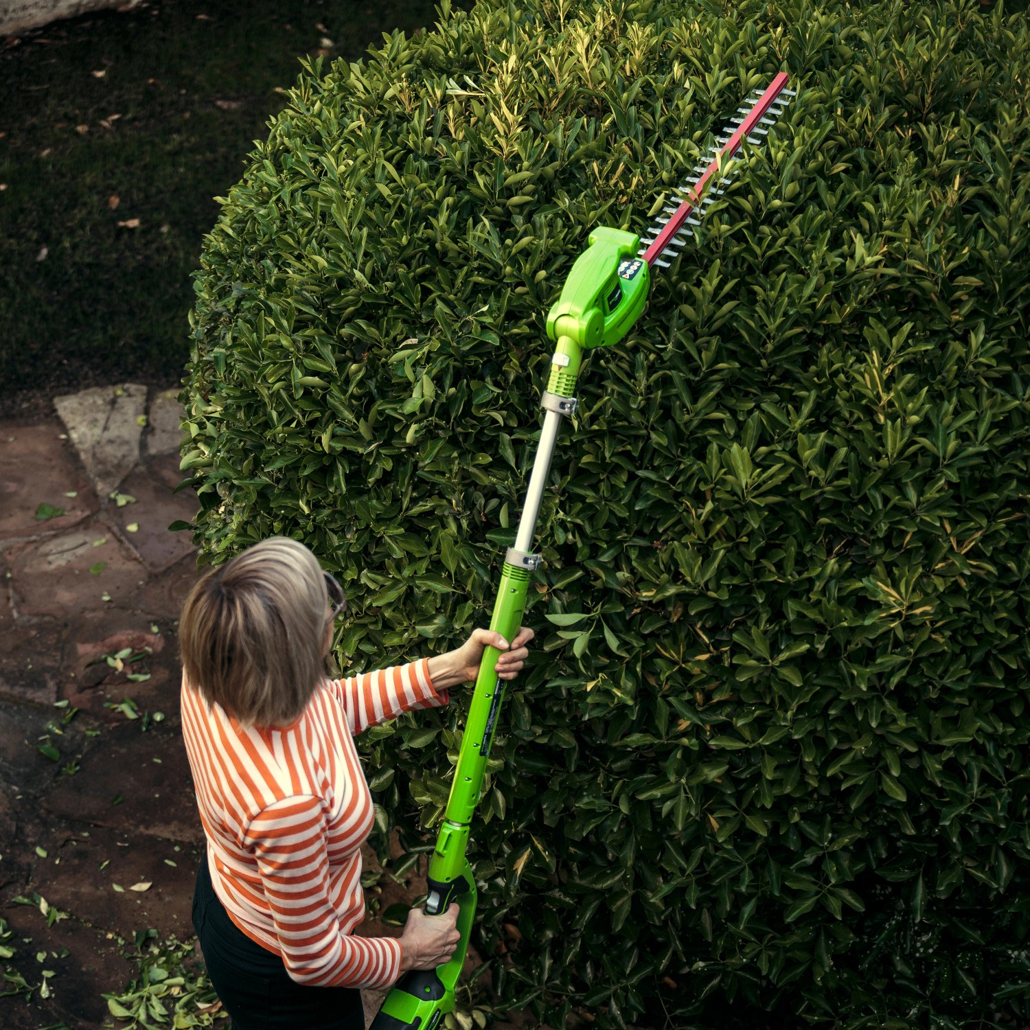 Frau schneidet hohe Hecke mit Greenworks Akku-Teleskop-Heckenschere G40PHA.