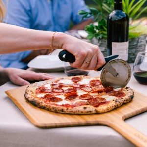 Ooni Pizzaroller schneidet Pizza mit Salami auf Holzbrett. Grillutensil mit Edelstahlklinge.