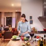 Szene mit Philips Hue E27 Lampe: Frau schneidet Gemüse in Küche, Wohnbereich mit warmem Licht im Hintergrund.