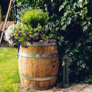 Dunkelgrüner Relaxdays Standbrunnen aus Aluminiumguss mit Wasserhahn neben bepflanztem Weinfass im Garten.