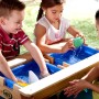 Kinder spielen am Sand- und Wasserspieltisch Nick Braun im Garten mit Wasser und Förmchen.
